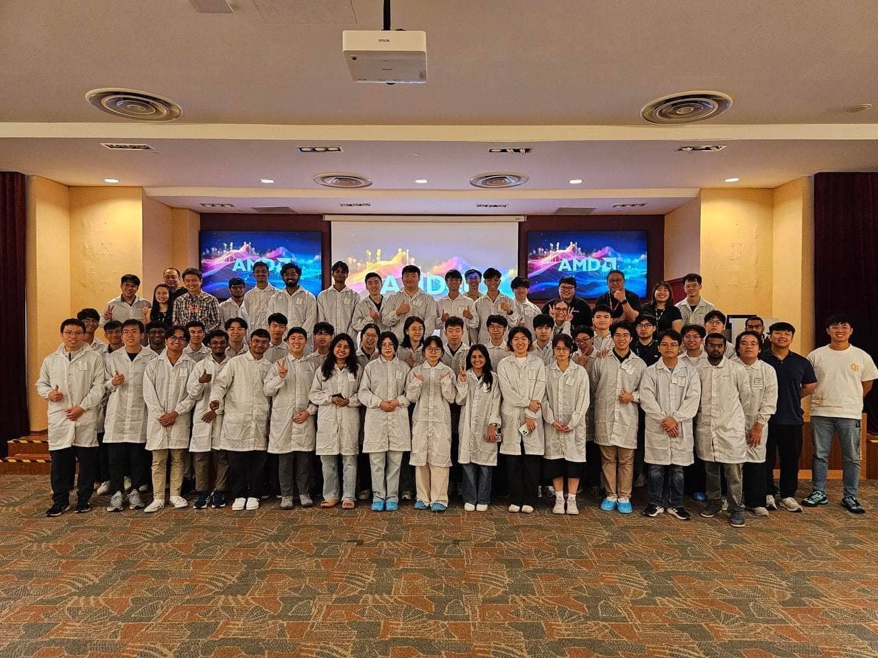 A large group of students in white coats posed in front of AMD screens during the site visit