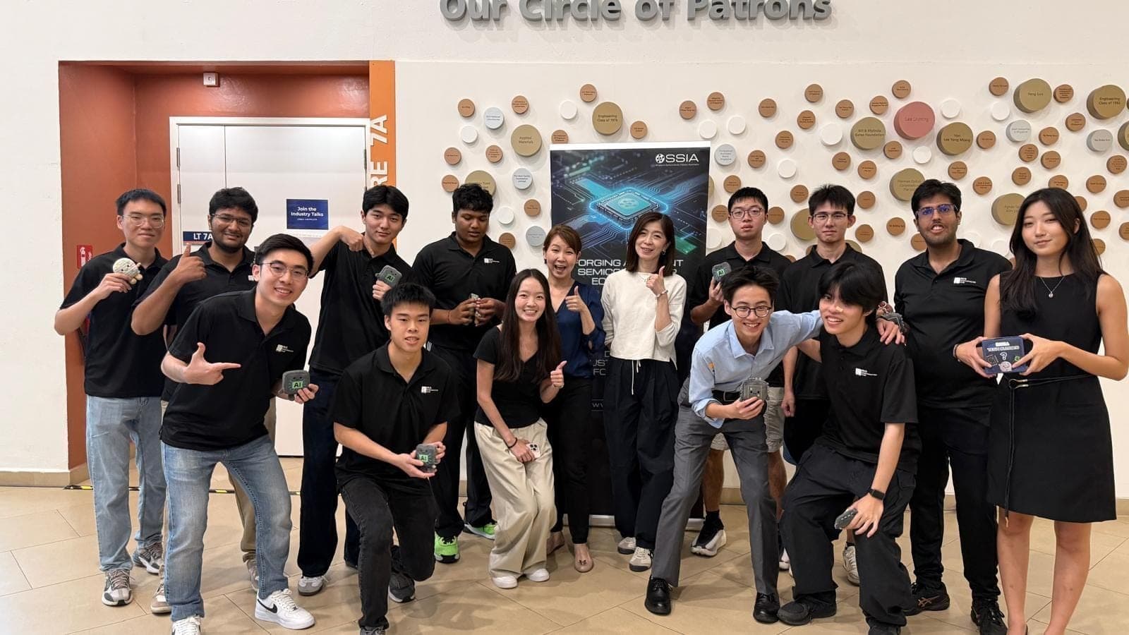 Club members in black polos with SSIA staff in front of an SSIA banner below the Circle of Patrons wall, several holding small gifts