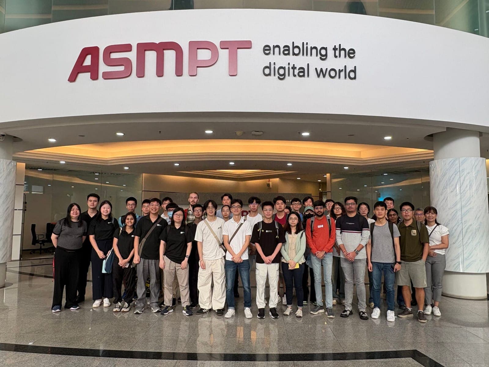 The visiting group photographed together beneath the ASMPT sign in the company's lobby